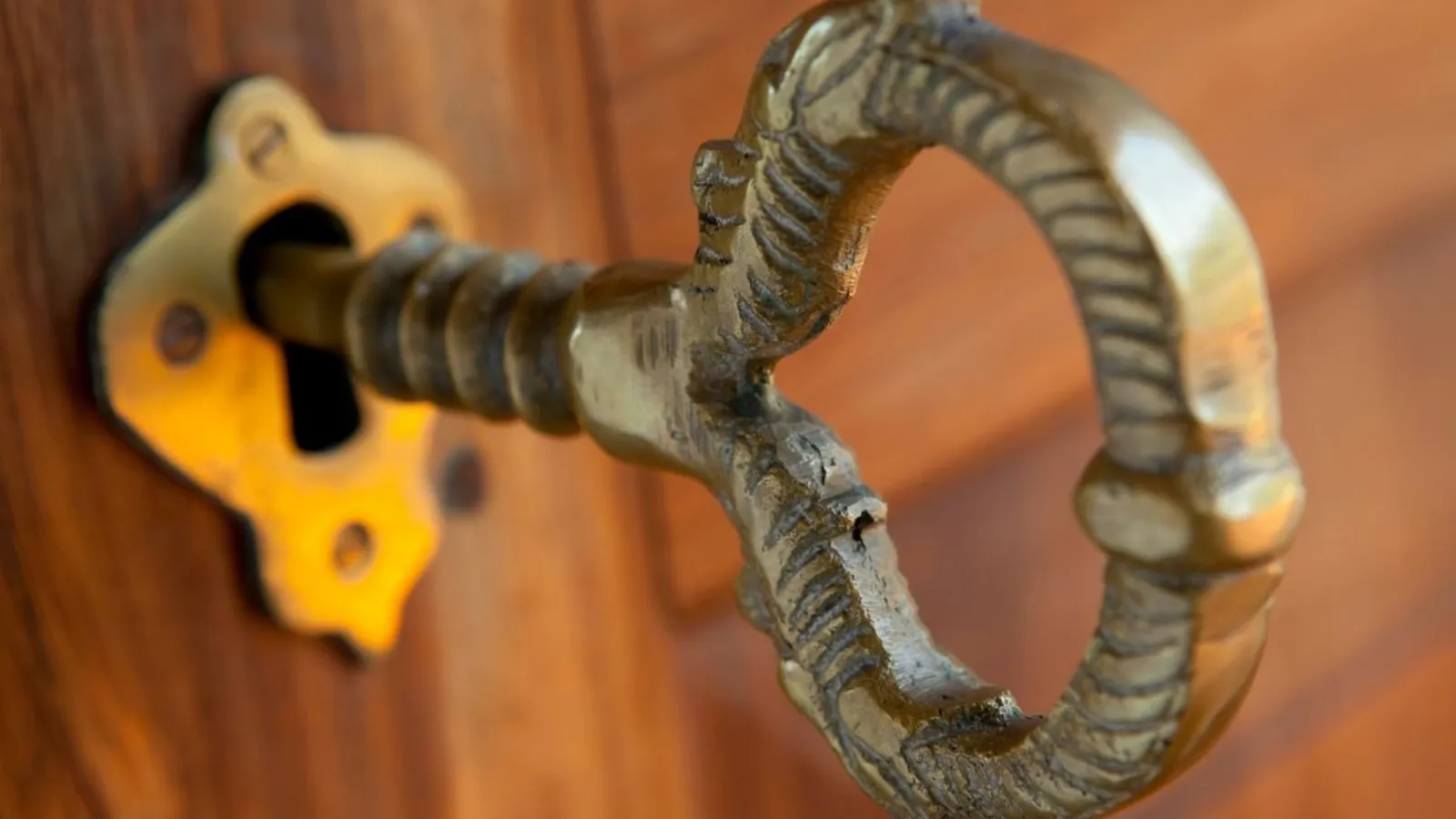 A close view of an ornate brass key left in the lock of a polished wooden door.