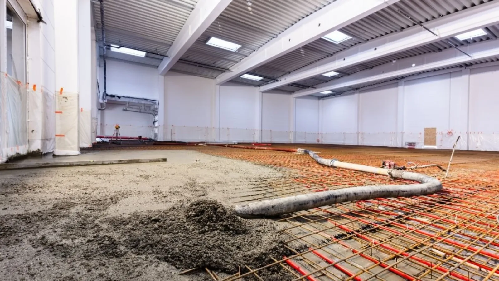 The inside of a large building under construction, reinforcing mesh and pipework laid bare across the floor with wet concrete poured over part of it.