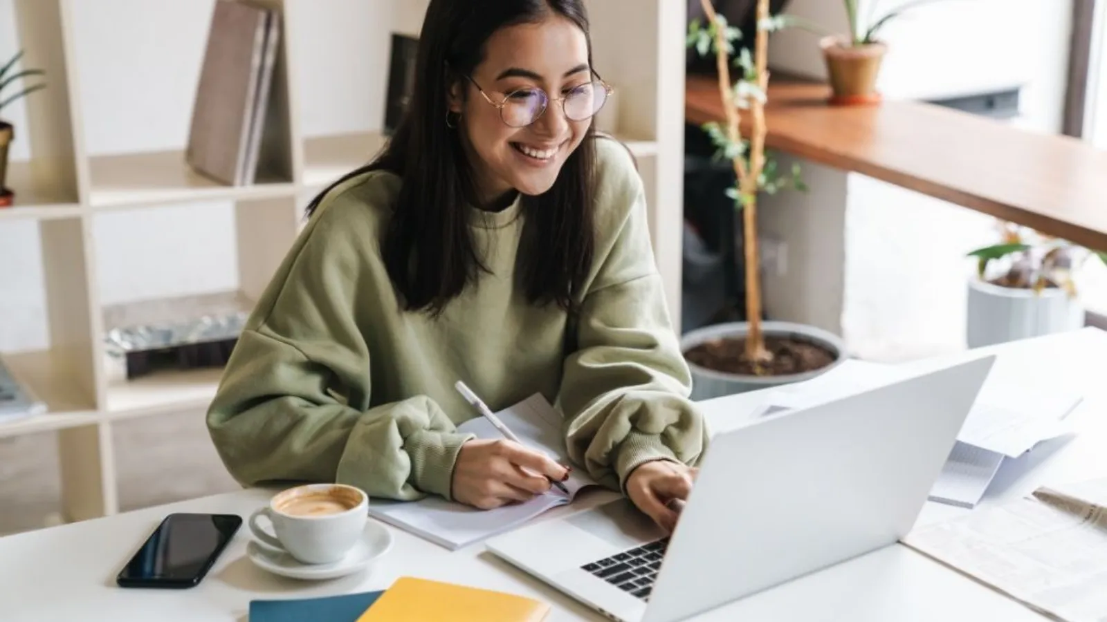 A smiling young woman in glasses writing in a notebook at a white desk, with a laptop, a cup of coffee and a phone beside her.