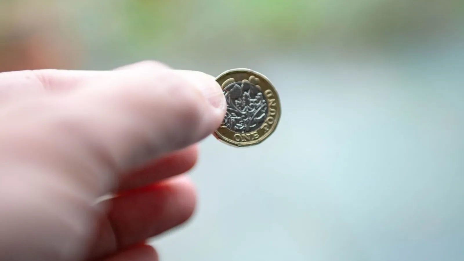 A hand holding a single British one pound coin between finger and thumb, against a blurred green and grey background.