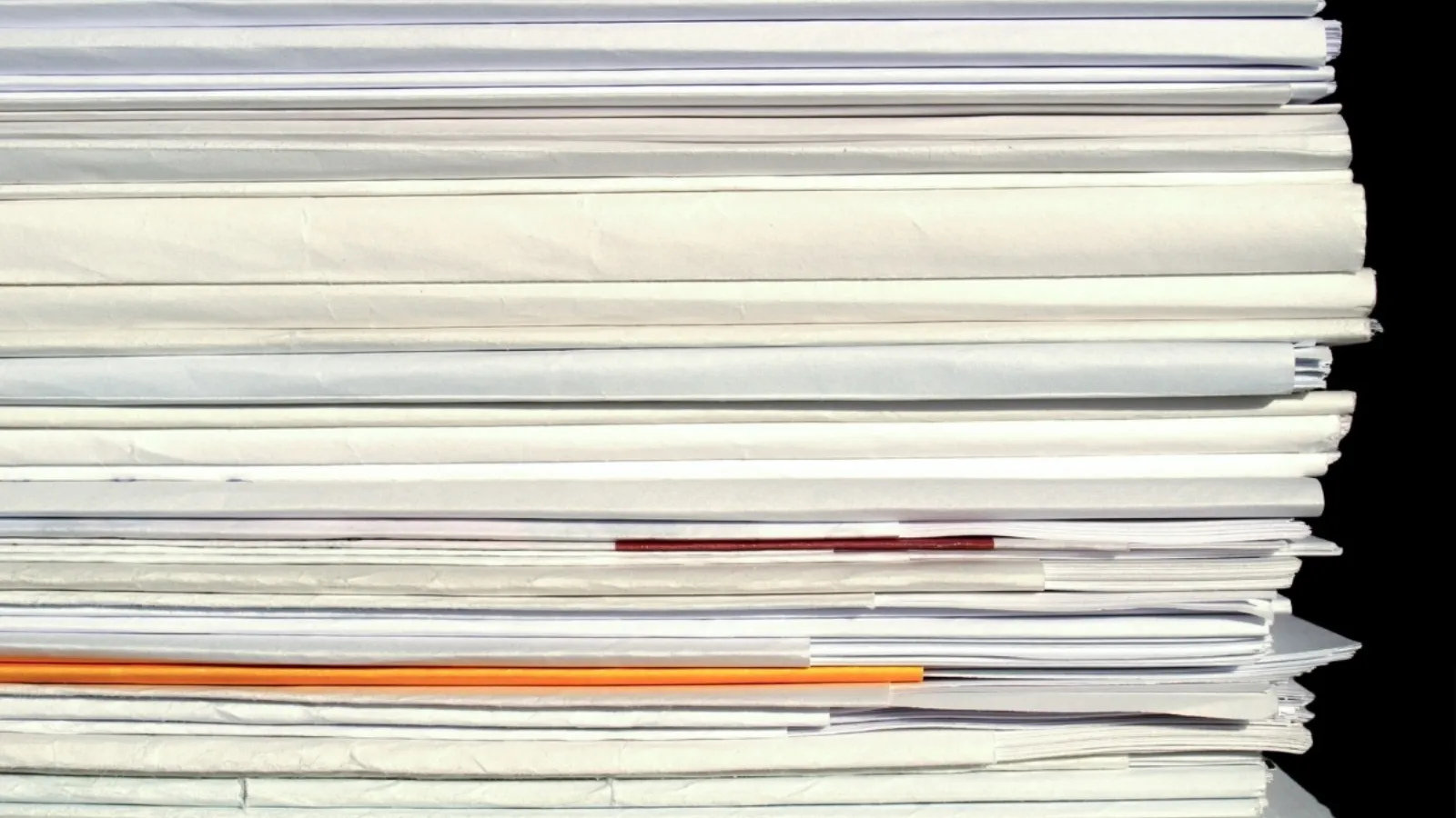 The edge of a thick stack of paperwork, a single orange sheet and one red-edged document among the white.