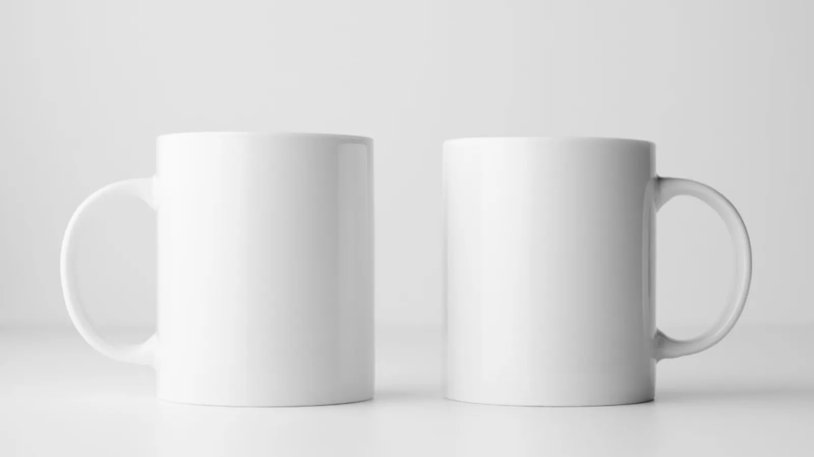Two plain white ceramic mugs side by side on a pale, evenly lit surface.