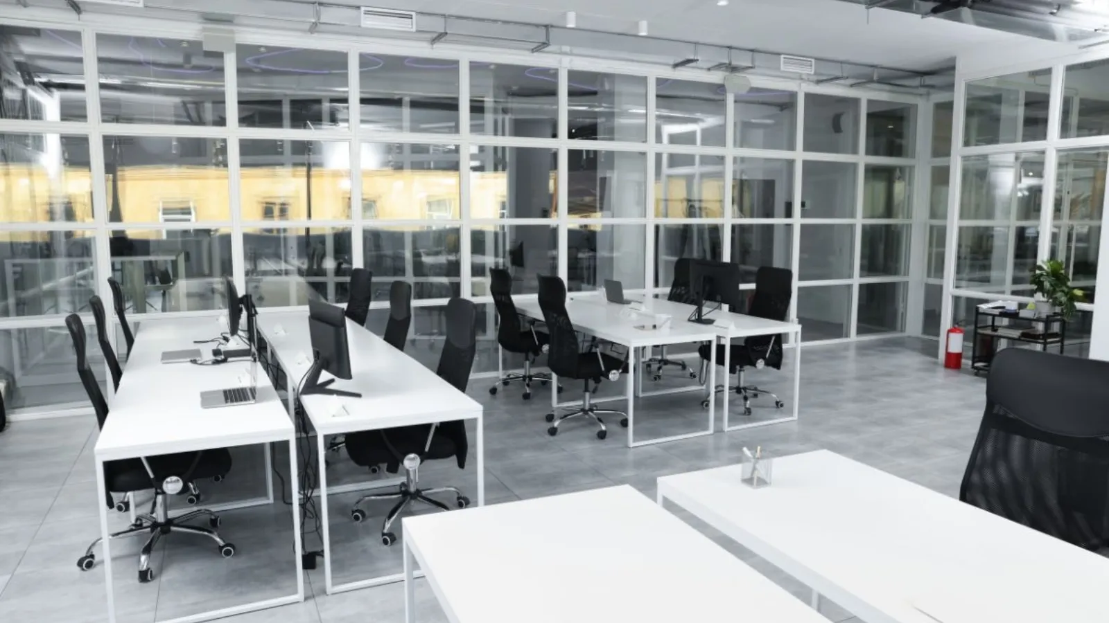 An empty open-plan office with white desks, black chairs and glass partitions, laptops and monitors left on the desks and nobody in the room.