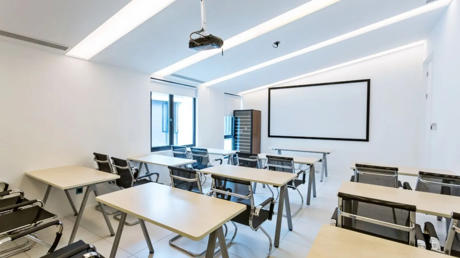 An empty training room with light wooden desks, black chairs and a blank screen at the front.